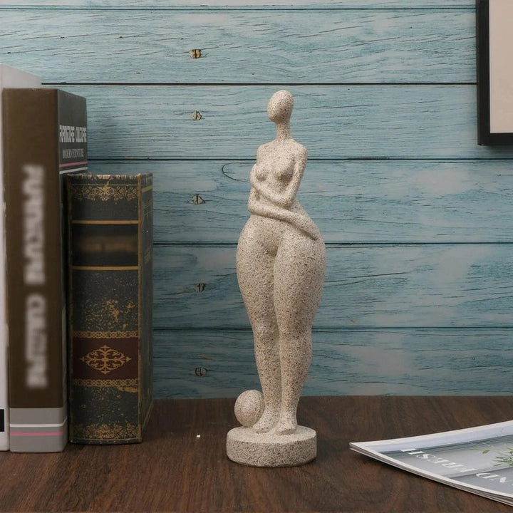 Statue of a nude figure on a wooden surface with books and a framed picture in the background.
