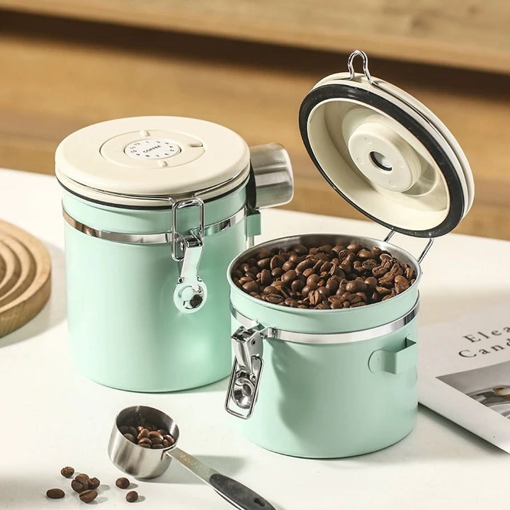 Mint green airtight coffee storage jars with cream lids, one opened showing coffee beans inside and a metal scoop beside.