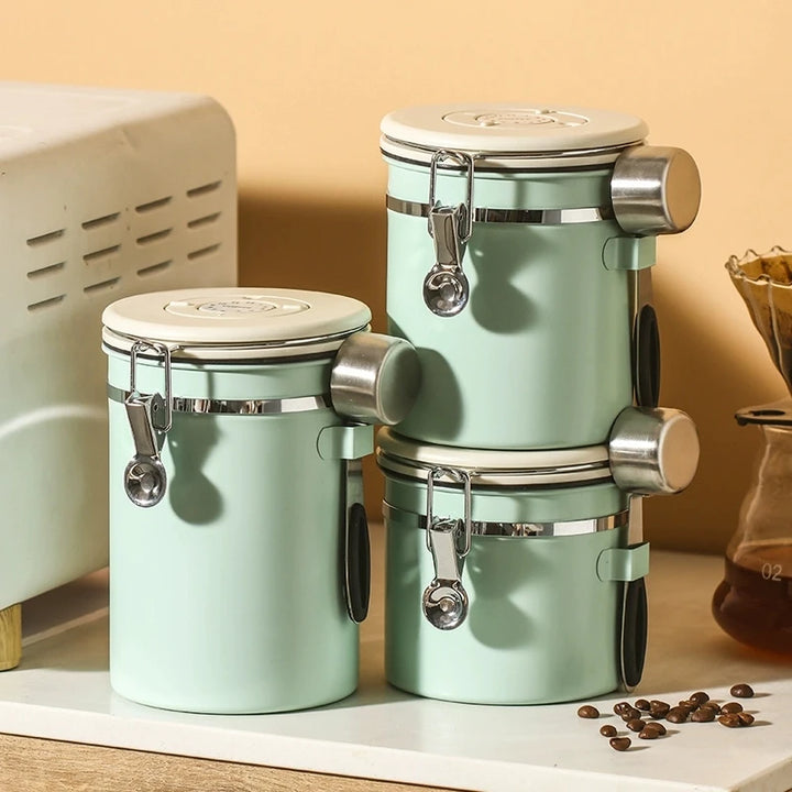 Stacked mint green stainless steel coffee storage jars with airtight seals and attached scoops on kitchen counter.