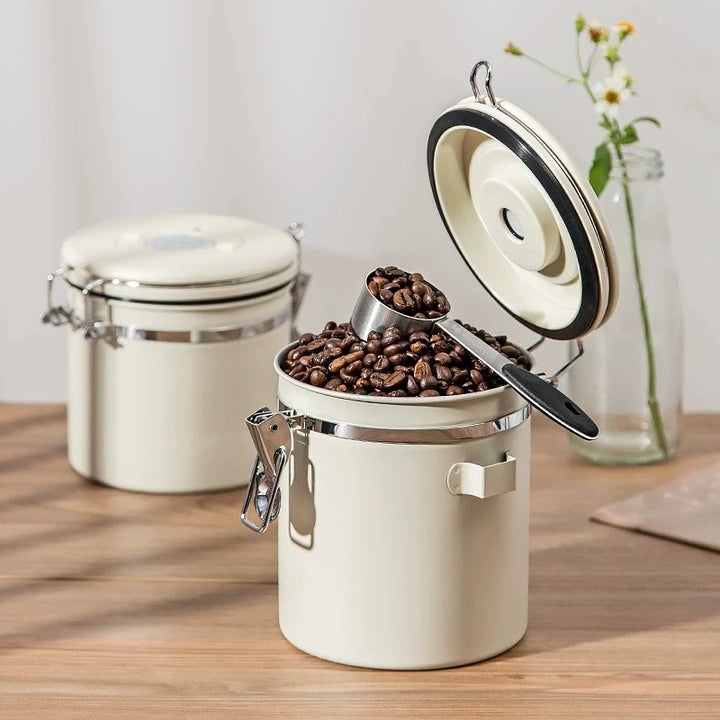 Open cream airtight coffee jar with metal scoop and coffee beans inside on a wooden table.