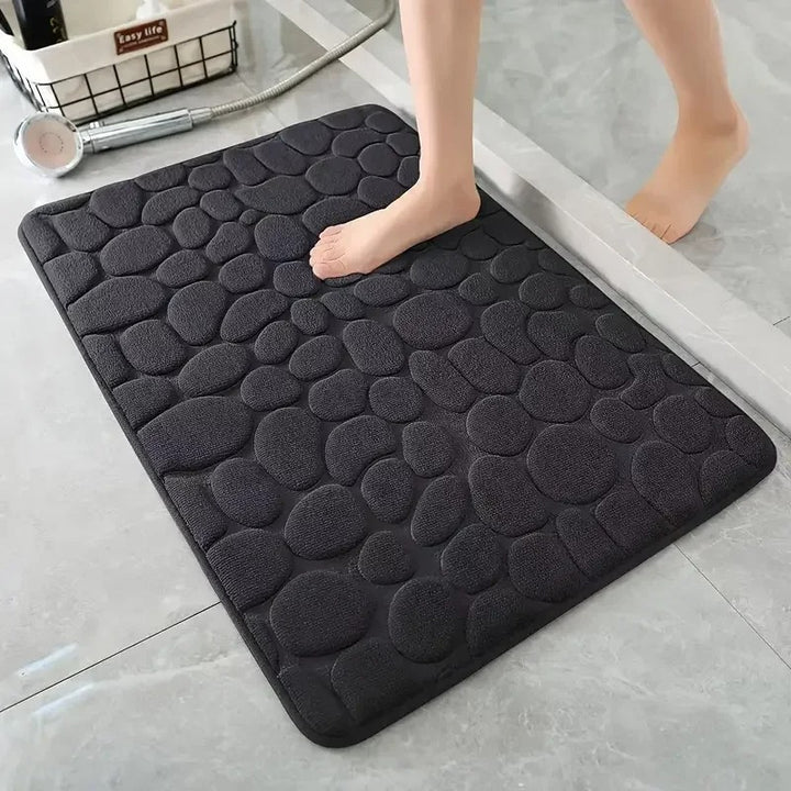 Black textured bath mat on a bathroom floor with a foot stepping onto it.