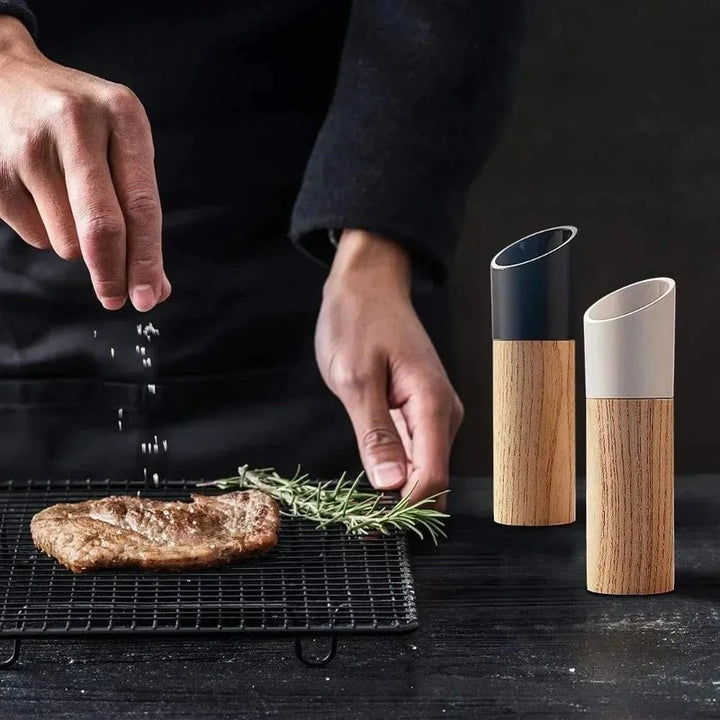 Person seasoning a piece of meat with salt and rosemary, next to two wooden salt and pepper shakers on a dark surface.