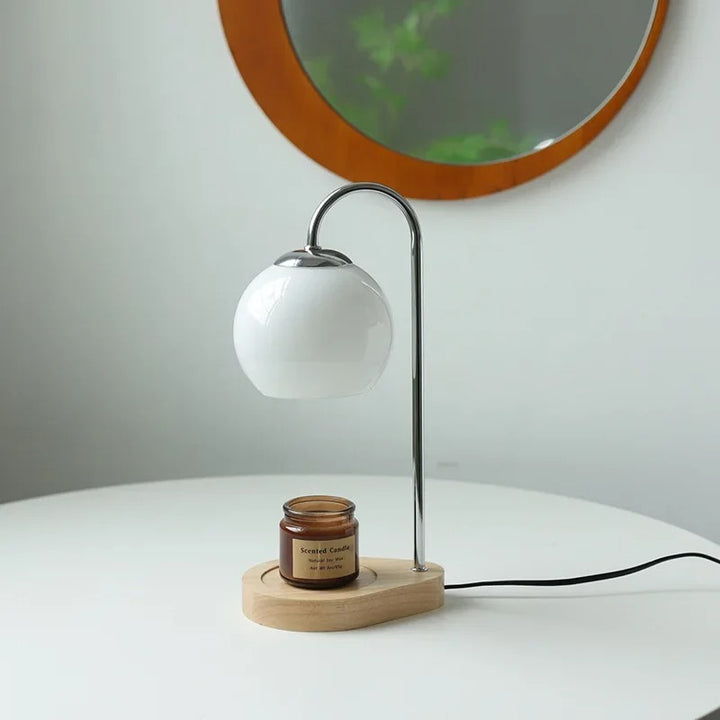 Table lamp with a white shade and wooden base on a white surface, with a mirror and plant in the background.