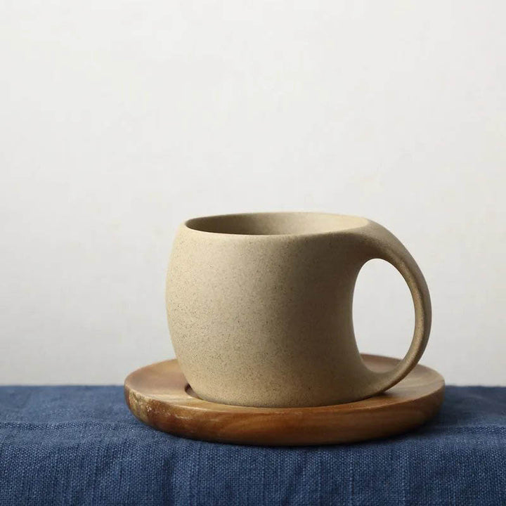 Beige ceramic mug with a smooth matte finish, paired with a natural acacia wood saucer on the blue fabric with white background.