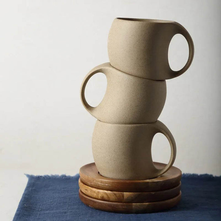 Stack of three ceramic mugs with a natural acacia wood saucer on the blue fabric with white background.