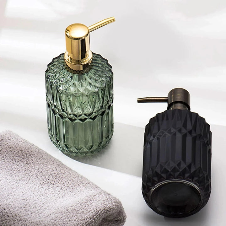 Two soap dispensers, one green with a gold pump and one black with a bronze pump, on a white surface with a towel.