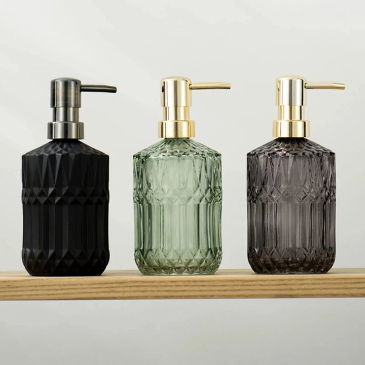 Three glass soap dispensers in black, green, and gray on a wooden shelf with a light background.