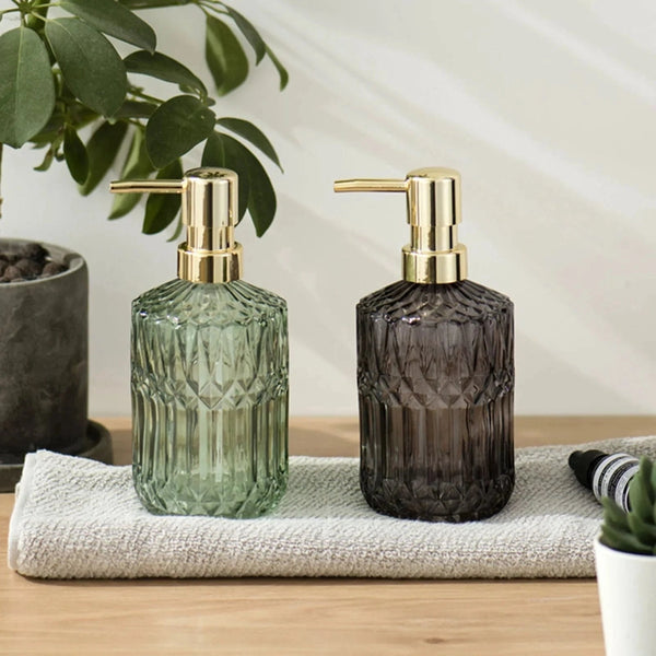 Two glass soap dispensers with gold pumps on a towel, surrounded by plants.