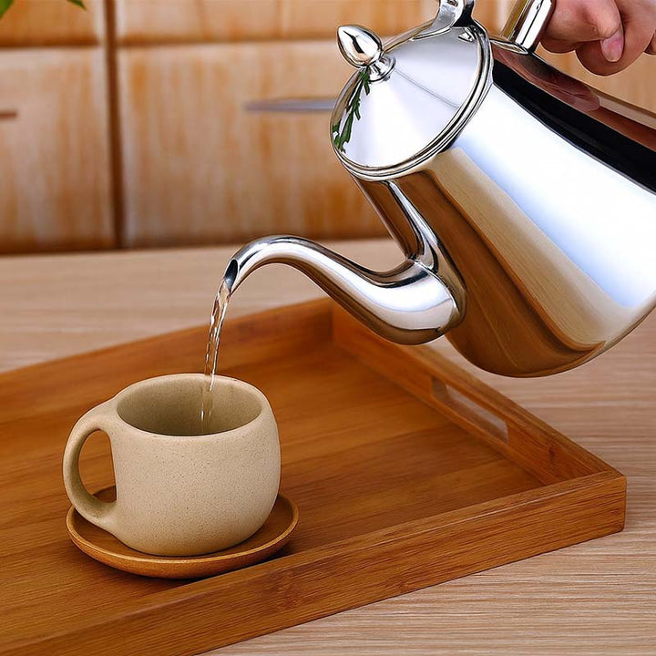 Silver stainless steel induction kettle pouring water into a beige mug on a wooden tray.
