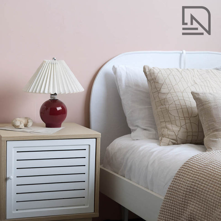 Red table lamp with pleated cream shade on white nightstand, cosy bedroom lighting for soft ambiance.