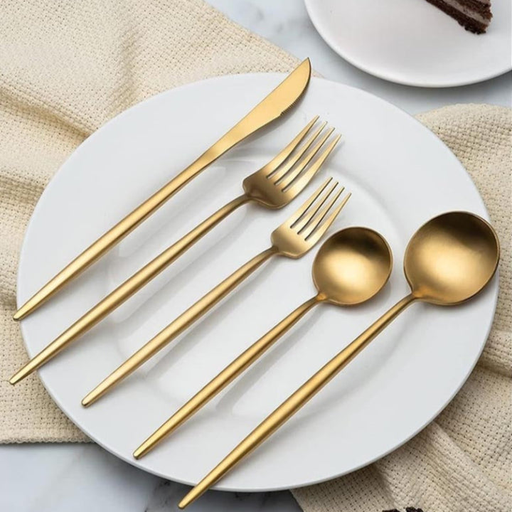 Gold 5-piece minimalist cutlery set on white plate: knife, two forks, teaspoon, soup spoon; modern tableware for dining.