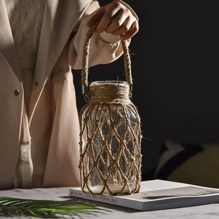 Hand holding a glass jar with a rope handle on a surface with a blurred background