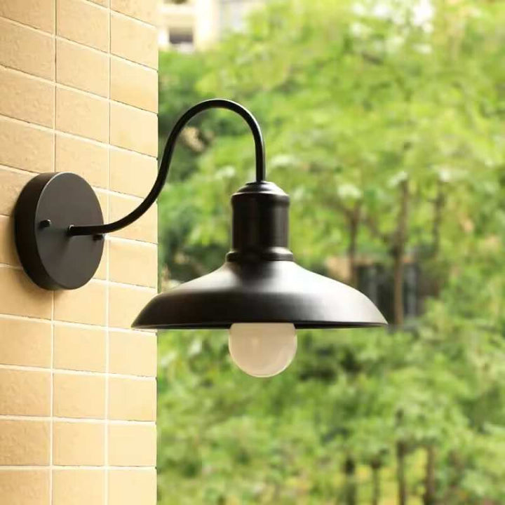 Matte black outdoor wall light with curved arm and LED bulb, mounted on beige tiled wall overlooking greenery