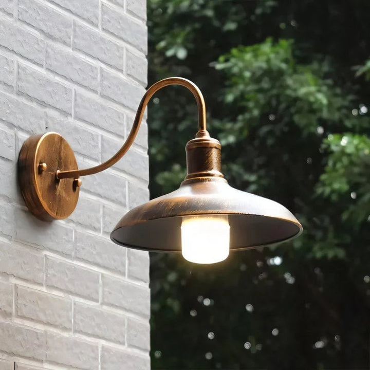 Vintage bronze outdoor wall lamp with curved arm and LED bulb, mounted on grey brick wall for garden or patio lighting