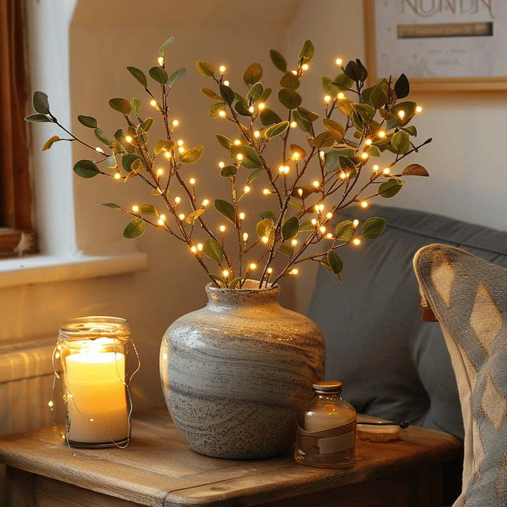 Fairy Light Olive Branch arrangement glowing warmly in a ceramic vase, styled beside a candle for a cozy living room atmosphere.