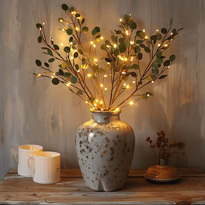 Warm glowing Olive Branch light arrangement in a rustic ceramic vase, styled on a wooden console with candles for cozy decor.