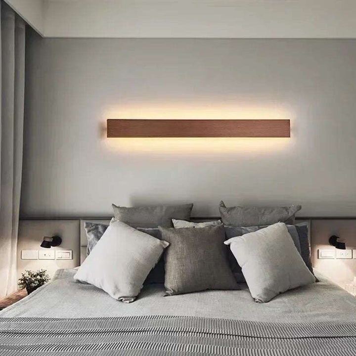 Long wooden finish LYST LED wall lamp above grey bed, creating warm Scandinavian bedroom lighting