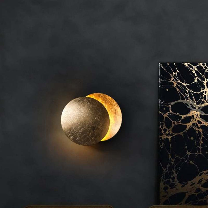 MÅNE Solar Eclipse Wall Light in brushed gold finish, mounted on a dark wall with artistic décor.