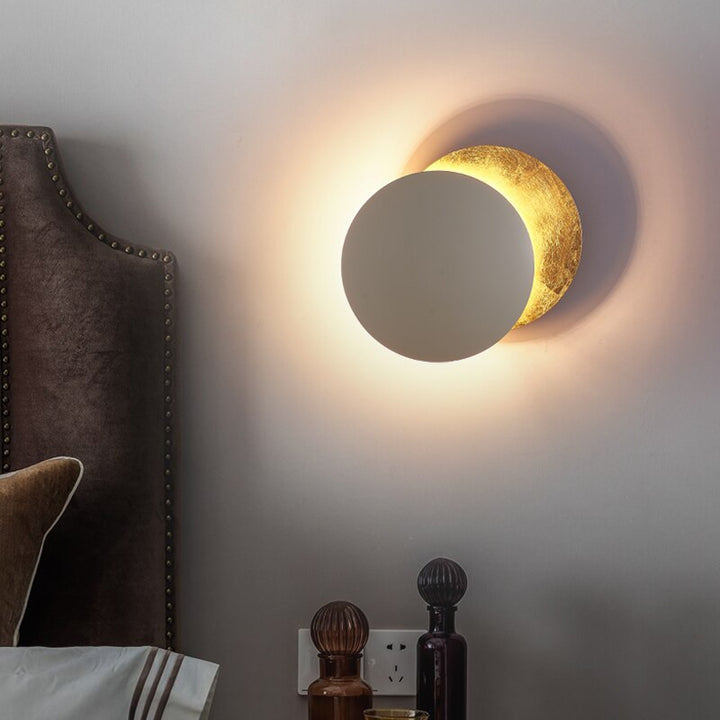 White and gold MÅNE Solar Eclipse Wall Light adding warmth beside a bed with soft ambient lighting.