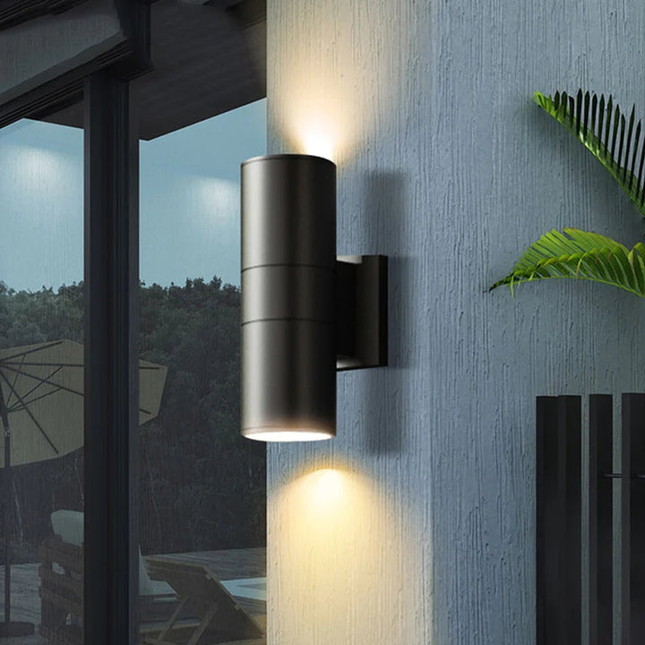 Black outdoor wall light with dual beam illumination mounted beside glass door and potted plant at night.