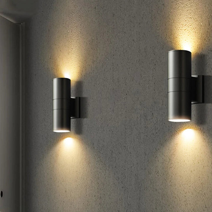 Pair of black cylindrical outdoor wall lights casting warm up and down illumination on textured grey exterior wall.