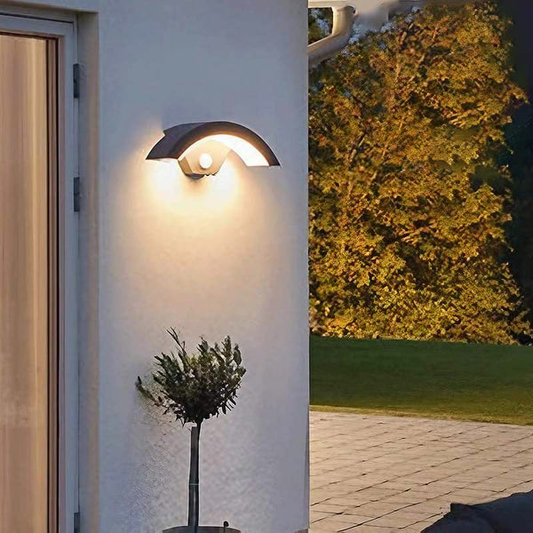Modern outdoor wall light fixture on a building exterior with garden and trees in the background.