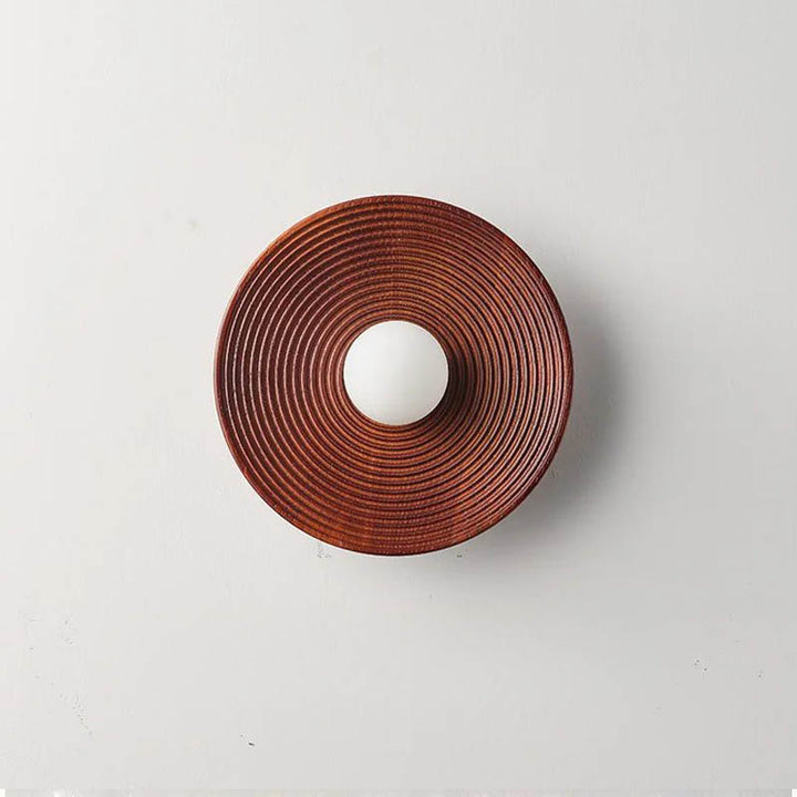Natural wood wall lamp in rich reddish-brown wood finish with centre glass diffuser, shown unlit against a neutral wall.