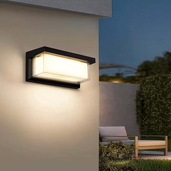 Modern outdoor wall light fixture on a house exterior with garden and bench in the background.