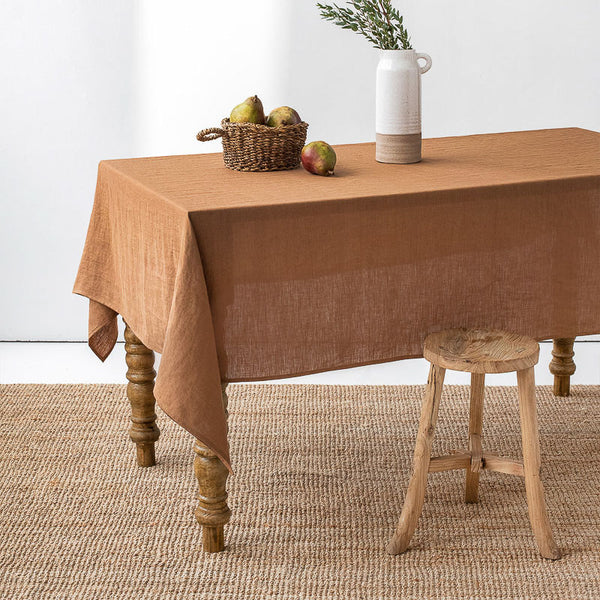 Brown Cinnamon linen tablecloth on a wooden table with a basket, apples, and a vase.