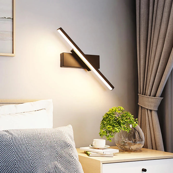 Modern rotatable wall-mounted light fixture in a bedroom setting with a plant and books on a nightstand.