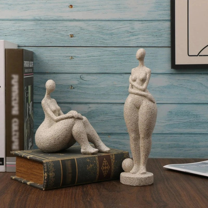Two stone sculptures of a sitting and standing figure on a wooden surface with a book and framed picture in the background.