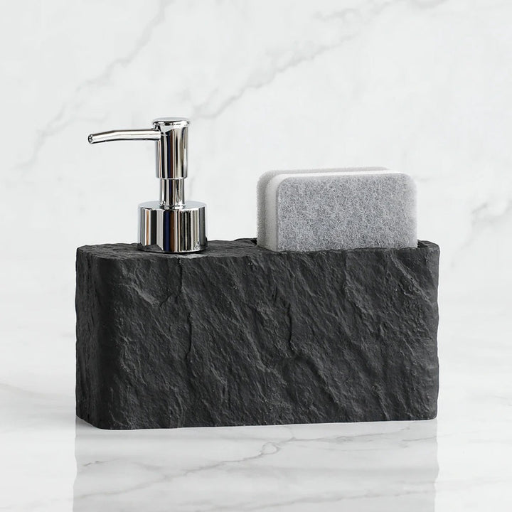 Sleek soap dispenser and sponge on a dark stone holder against a white marble background
