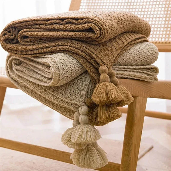 Neutral-toned Nordic knitted throw blankets with tassels, soft cotton blend, folded on chair, ideal for sofa or bed.