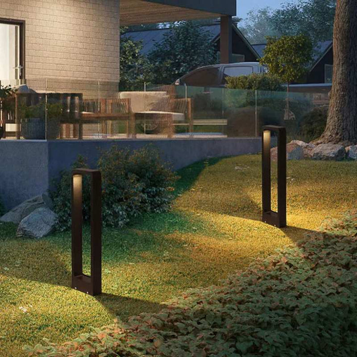 Two outdoor garden lights illuminating a grassy area with a patio and trees in the background.
