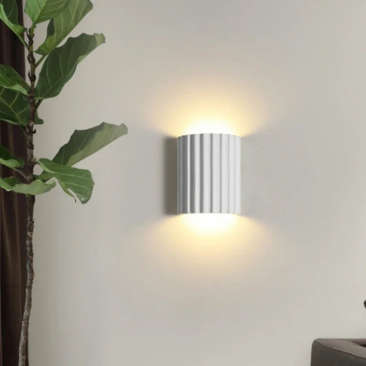 Wall-mounted light fixture on a white wall with a plant to the left.