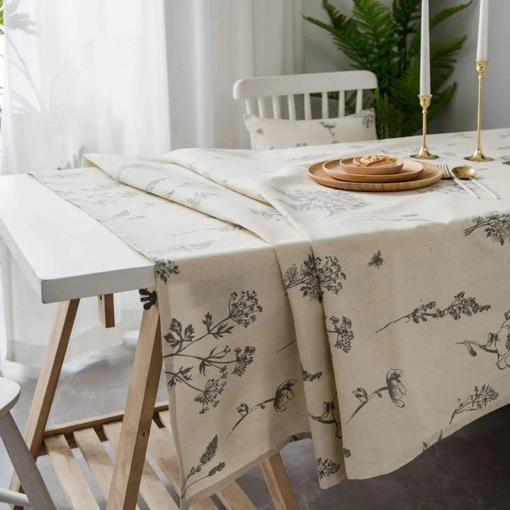 Dining table set with a floral-patterned tablecloth, plates, and candles in a bright room.