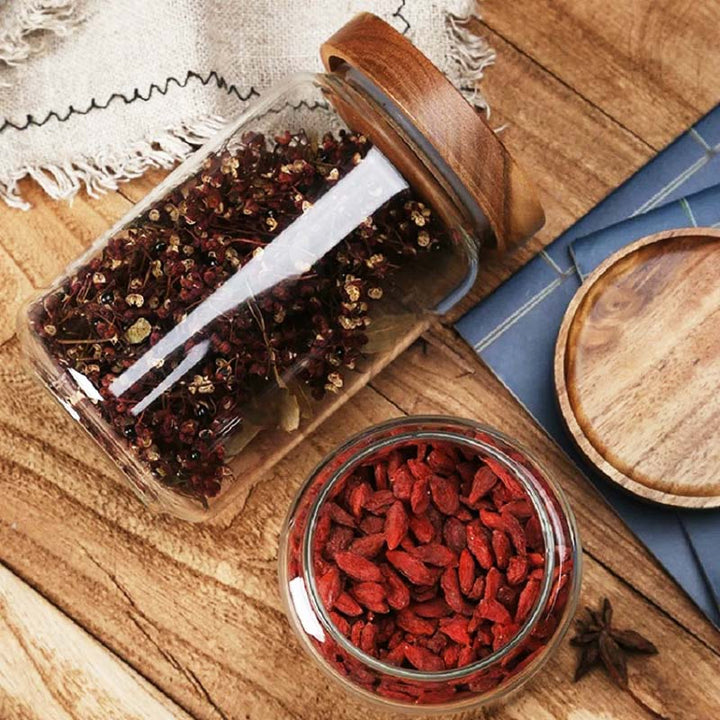 Two glass jars on a wooden surface, one with red dried berries and the other with whole spices.